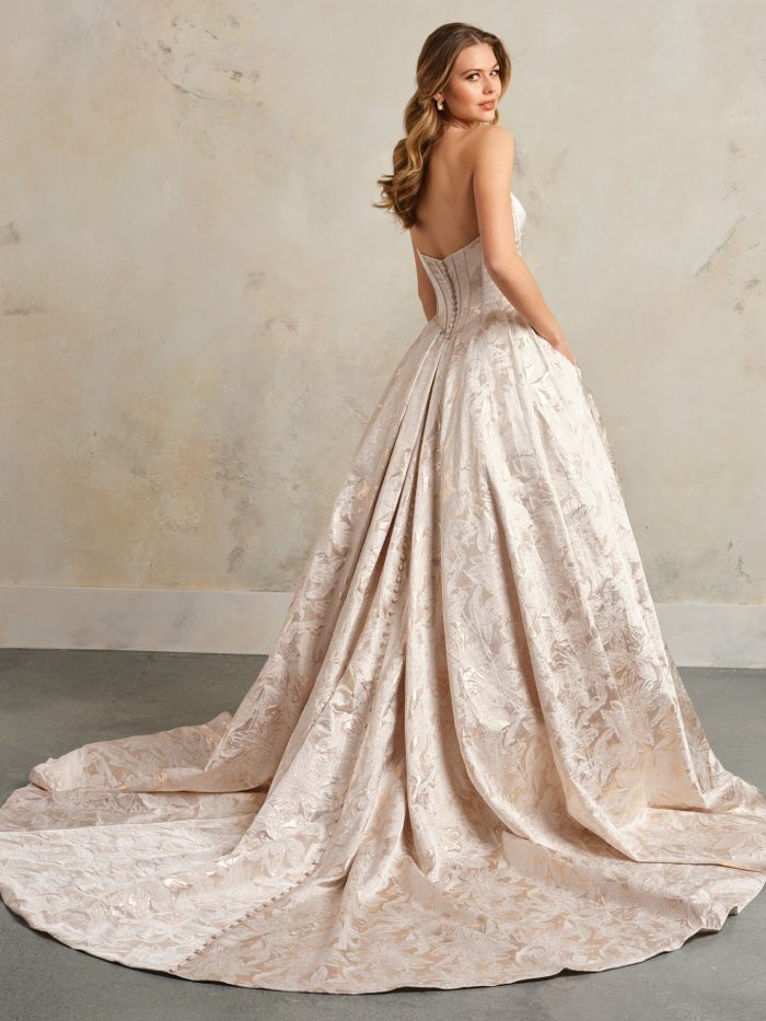 ack view of the Derrick Marie ball gown wedding dress by Maggie Sottero, showing a low sweetheart back with covered button closure down the full metallic floral jacquard skirt and train, in a blush colorway.