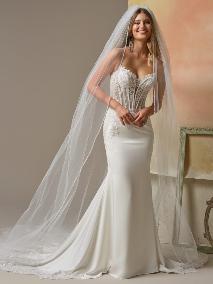 Maggie Sottero India Sheath wedding dress in white, front view featuring a sweetheart sheer lace corset bodice with 3D floral appliqué and beaded spaghetti straps, smooth crepe sheath skirt with lace hip detail, and cathedral train, styled with a crystal-edged cathedral veil.