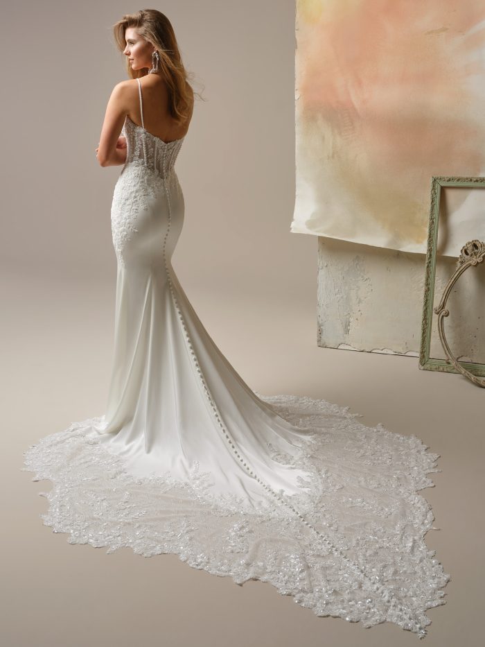 Maggie Sottero India Sheath wedding dress in white, back view showing sheer illusion lace back with spaghetti straps and covered button closure, smooth crepe sheath skirt, and dramatic cathedral train with ornate beaded lace border.