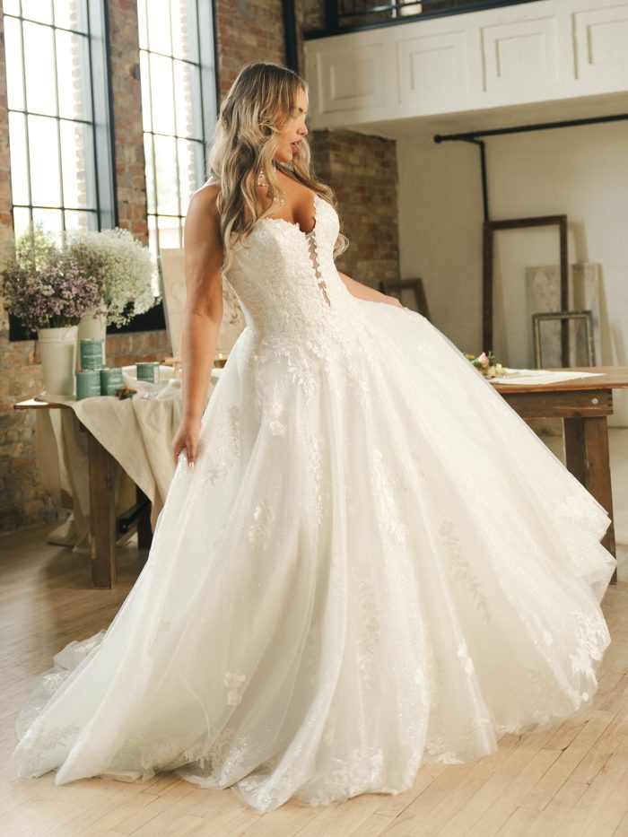 Maggie Sottero Jennifer ballgown wedding dress in ivory, curve fit — strapless plunging sweetheart neckline with lace bodice over a full glittering tulle ballgown skirt with floral appliqués, styled in a bright studio.