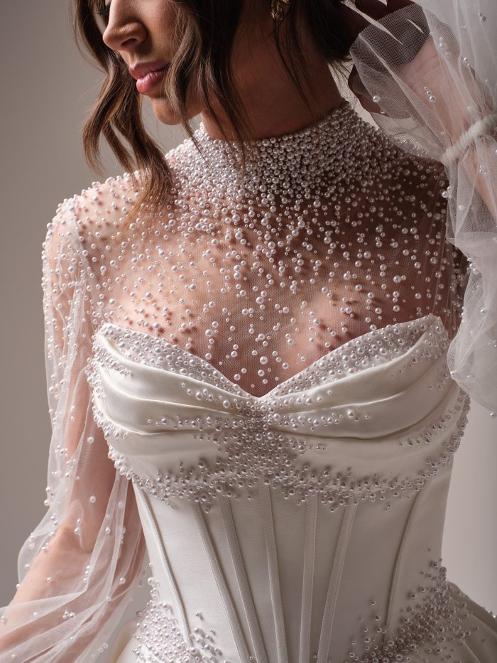 Close-up of a bride wearing the Juneau gown by Maggie Sottero, showing a fully pearl-beaded high neckline with sheer long sleeves and a draped satin sweetheart bodice.