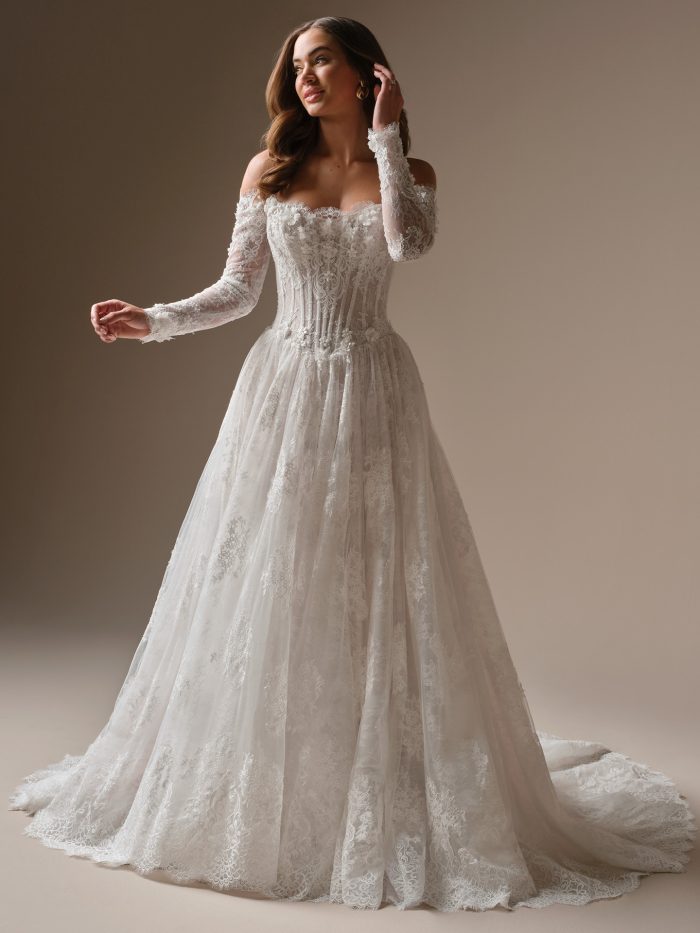 Bride wearing the Kitaro ballgown wedding dress, featuring an off-the-shoulder lace sweetheart neckline, sheer long lace sleeves with floral appliqué, a corset bodice with visible boning, and a full lace ballgown skirt with a scalloped hem.