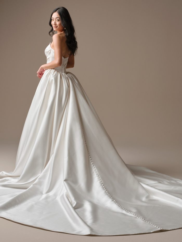 Maggie Sottero Maureen Lane Fit and Flare wedding dress in white, back view showing open sweetheart back with covered button closure, voluminous smooth satin skirt, and dramatic cathedral train with covered button detail along the edge.