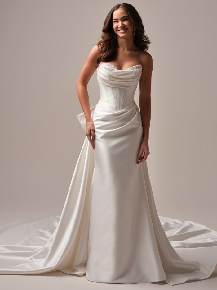 A bride wears the Melissa gown by Maggie Sottero, a satin fit-and-flare dress with a draped cowl neckline, fitted corset bodice, and a detachable bow at the hip.