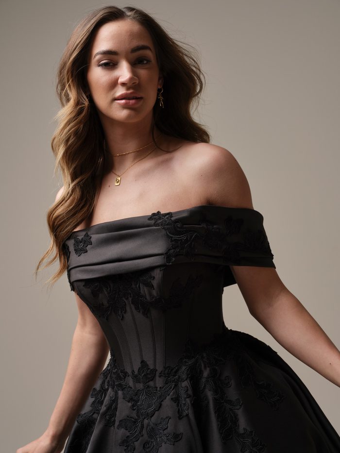 Close-up of a model wearing the Serafina gown by Maggie Sottero in black, showing an off-the-shoulder folded neckline with floral lace appliqué on the bodice.