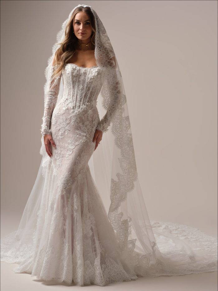 Maggie Sottero Takera Fit and Flare wedding dress in ivory, front view featuring a strapless sweetheart sheer lace corset bodice with 3D floral detail, long sheer lace sleeves, body-skimming lace fit and flare skirt, and cathedral train, styled with a matching lace-edged cathedral veil.