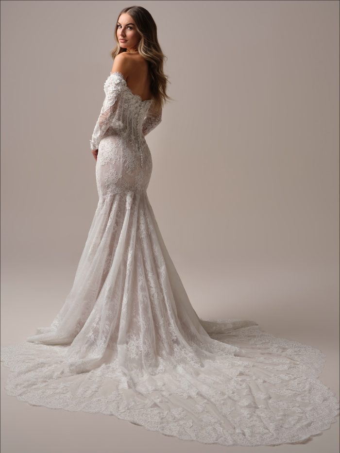 Maggie Sottero Takera Fit and Flare wedding dress in ivory, back view showing open low back with covered button closure, detachable off-the-shoulder long sheer lace sleeves, body-skimming all-over lace silhouette, and sweeping cathedral train with scalloped lace border.