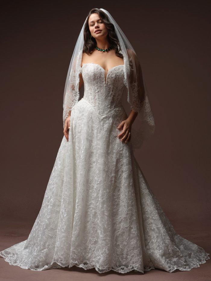 Maggie Sottero Zahia A-line wedding dress in ivory, curve fit — off-the-shoulder plunging sweetheart neckline with lace sleeves, fitted lace bodice, full A-line skirt, styled with a lace-trimmed veil.