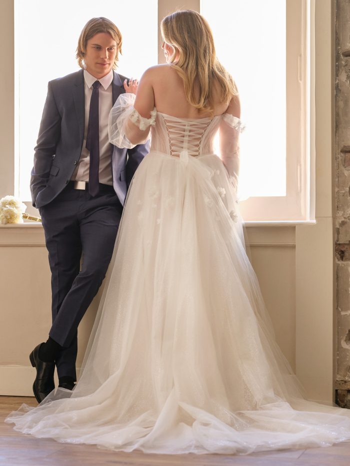Rebecca Ingram Allison A-line wedding dress in ivory, curve fit — back view showing off-the-shoulder sheer sleeves, corset lace-up back, and full glittery tulle skirt with 3D floral appliqués, styled with a groom by a window.