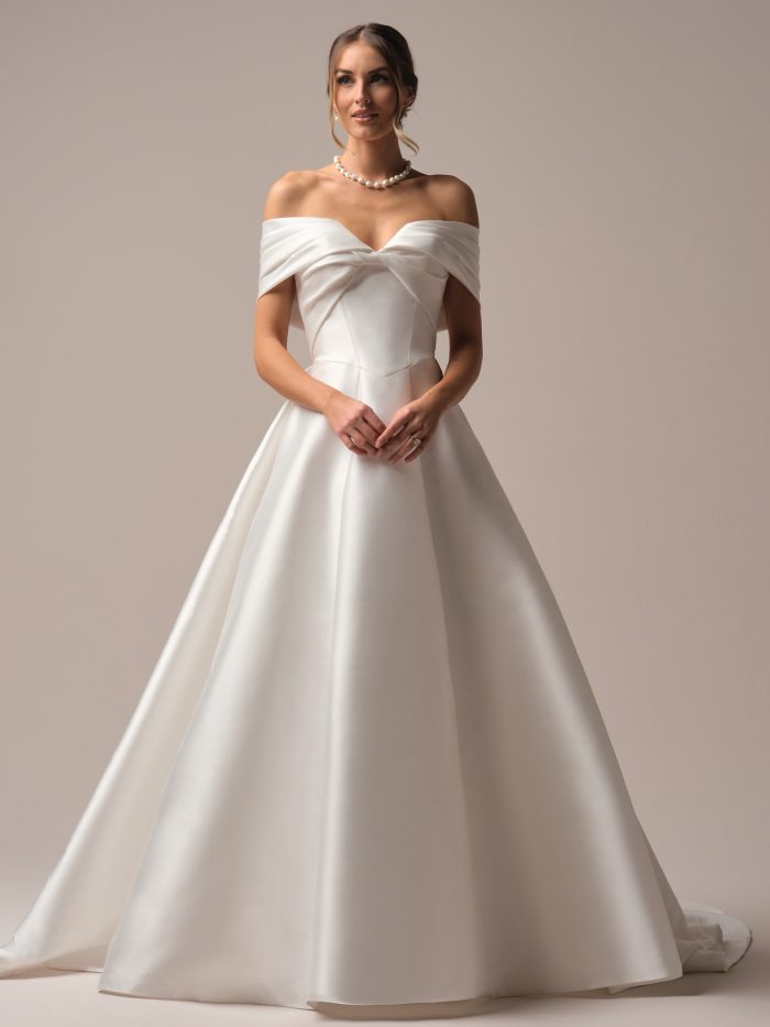 A bride wears the Brooke gown by Rebecca Ingram, a satin ball gown with an off-the-shoulder folded neckline and a full pleated skirt, paired with a pearl necklace.