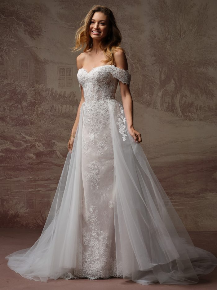 A bride wears the Esmeralda Lane gown by Rebecca Ingram, a lace fit-and-flare dress with an off-the-shoulder sweetheart neckline and a sheer tulle overskirt.