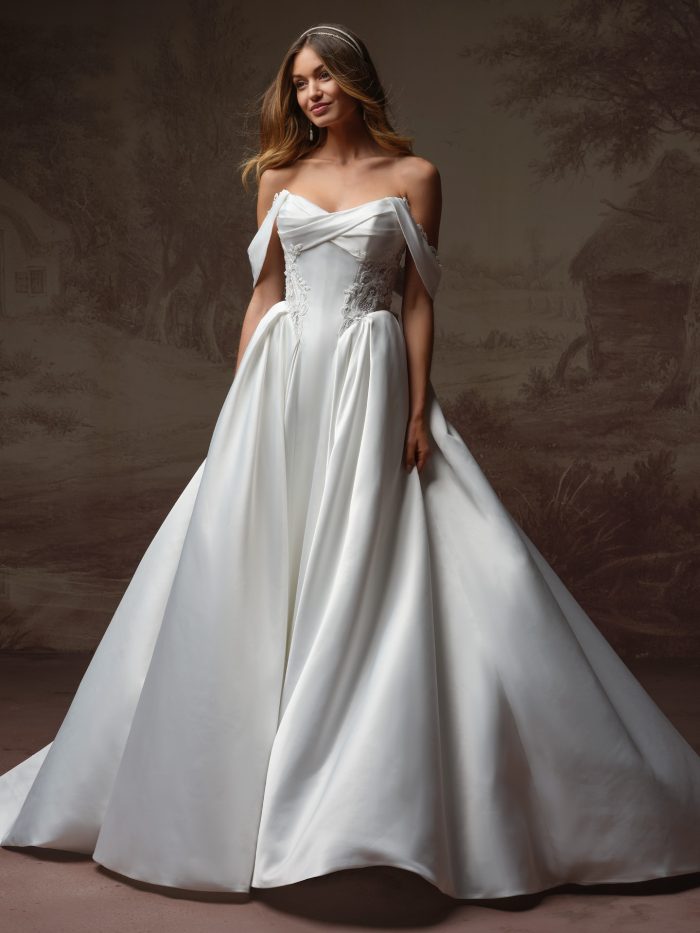 A bride wears the Gigi gown by Rebecca Ingram, a satin ball gown with an off-the-shoulder sweetheart neckline, crisscross draped bodice, and a voluminous pleated skirt with side pockets.
