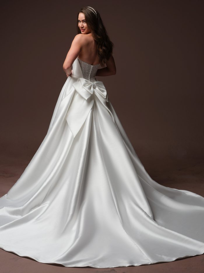 Back view of a bride wearing the Joanie gown by Rebecca Ingram, a satin ball gown with a beaded corset bodice and a large satin bow at the back waist with a full train.