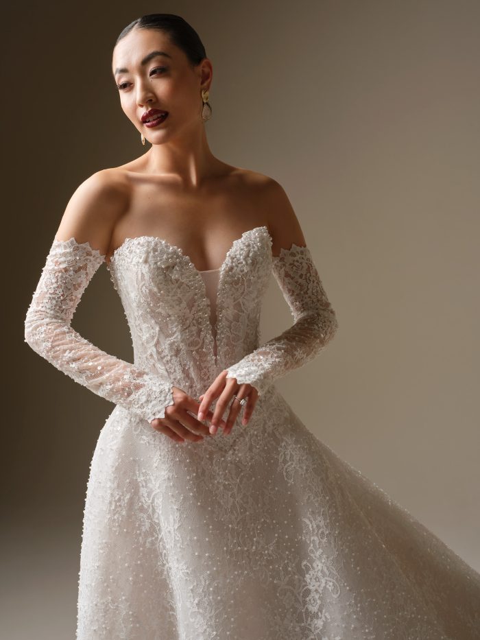 Close-up of the Angelica gown by Sottero and Midgley, highlighting the beaded lace off-the-shoulder neckline and sheer embellished sleeves.