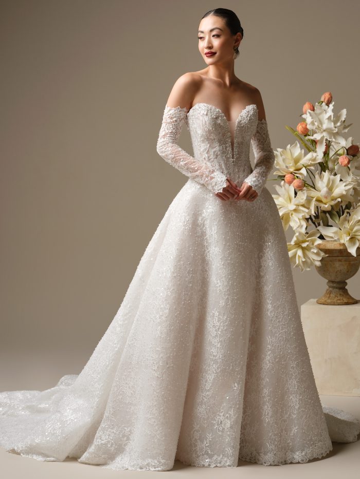 A bride wears the Angelica gown by Sottero and Midgley, a sparkling lace A-line dress with an off-the-shoulder sweetheart neckline, sheer beaded long sleeves, and a full train, standing beside a floral arrangement.