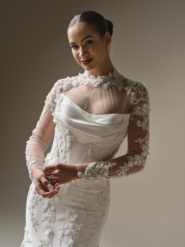 A bride wears the Burke gown by Sottero and Midgley, a fitted dress with a high sheer lace neckline, draped satin bodice detail, and long floral lace sleeves.