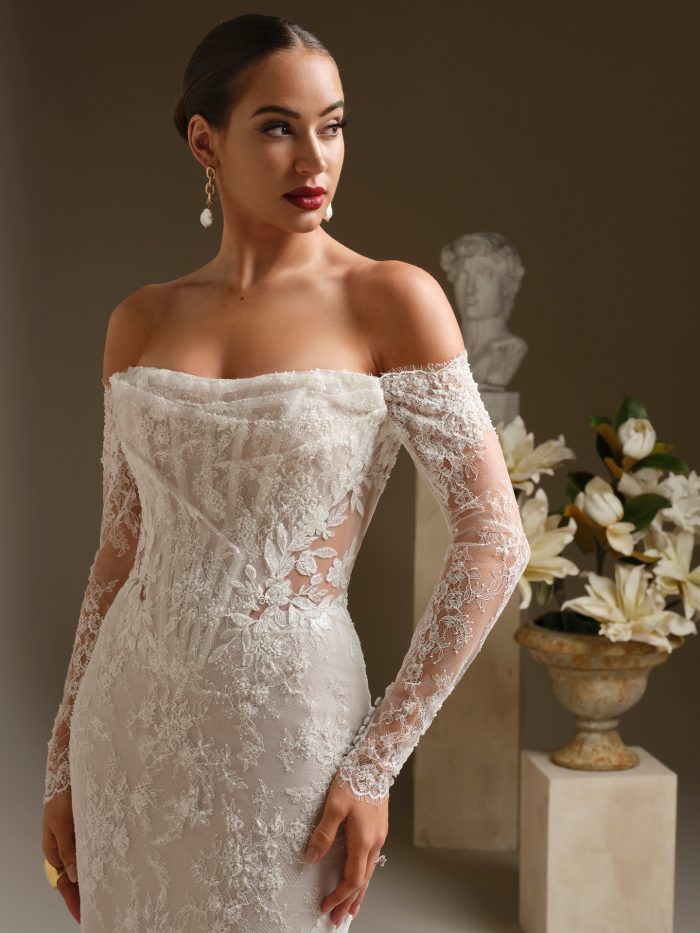 A bride wears the Dublin gown by Sottero and Midgley, a fitted lace sheath dress with an off-the-shoulder draped neckline and sheer floral lace long sleeves.