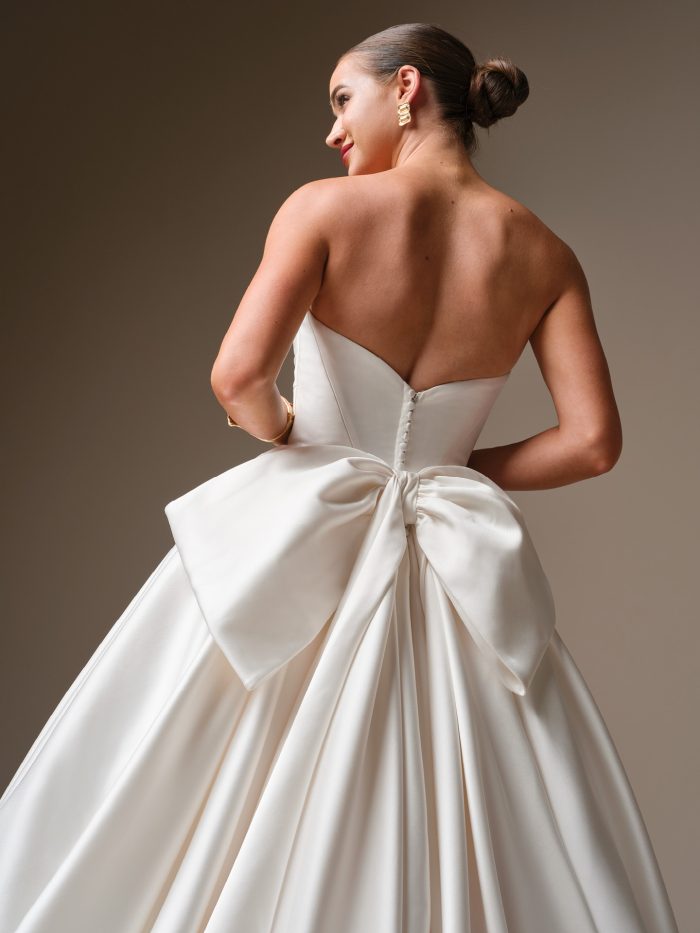 Back view of a bride wearing the Elias gown by Sottero and Midgley, a satin ball gown with a strapless bodice, covered buttons, and an asymmetrical satin bow at the back waist.