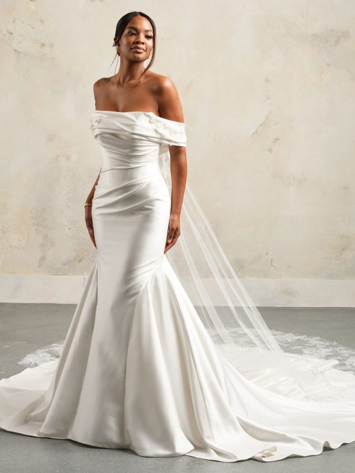 A bride wears the Giselle gown by Sottero and Midgley, a fitted satin mermaid dress with an off-the-shoulder draped neckline and a long lace-trimmed veil.
