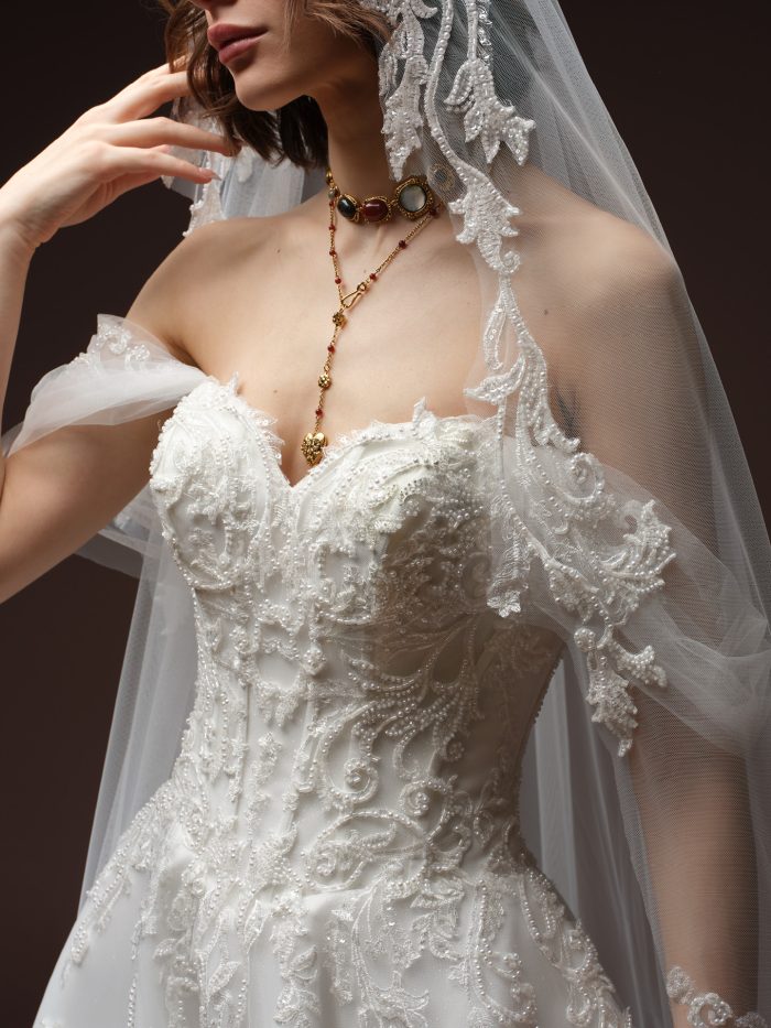 Close-up of a bride wearing the Gwyneth gown by Sottero and Midgley, showing the beaded lace bodice with sweetheart neckline, sheer puff sleeve, lace-edged veil, and layered gold jeweled necklace.