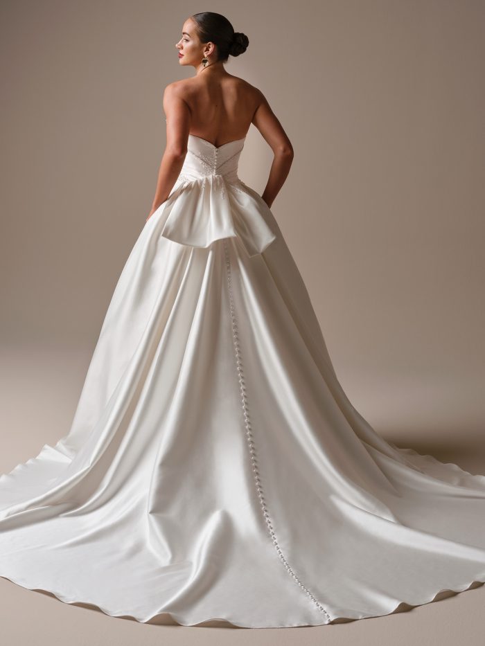 Sottero and Midgley Kincaid Ballgown wedding dress in white, back view showing open sweetheart back with large satin bow detail, covered button closure running the length of the cathedral train, and voluminous smooth satin skirt with sweeping cathedral train.
