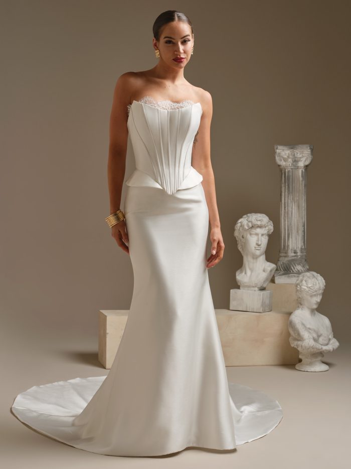 A bride wears the Lassen Marie gown by Sottero and Midgley, a fitted satin dress with a strapless corset bodice, lace trim at the neckline, and a slim skirt with a train, standing beside classical statues.