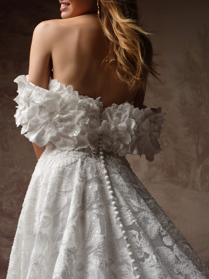 Back view of the Merrigan gown by Sottero and Midgley, showing the ruffled off-shoulder detail, covered buttons, and full lace skirt.