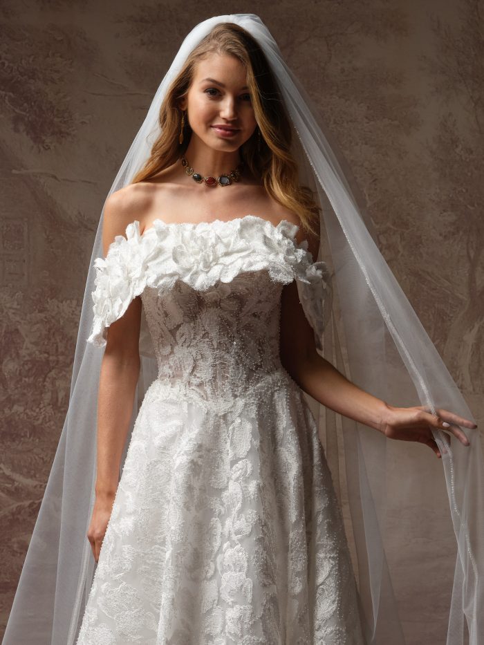 A bride wears the Merrigan gown by Sottero and Midgley, a lace A-line dress with an off-the-shoulder ruffled floral neckline, paired with a flowing veil and jeweled necklace.