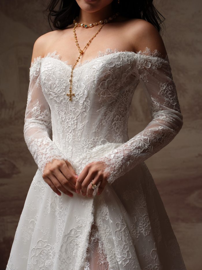 Close-up of the Vespera wedding dress, featuring an off-the-shoulder lace sweetheart neckline with sheer floral lace long sleeves and a fitted lace bodice.