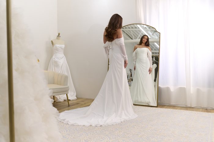 Bride admiring her reflection in a boutique mirror while wearing the Francesca gown by Rebecca Ingram, a chiffon off-the-shoulder dress with a flowing train.