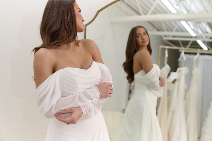 Bride looking over her shoulder at her reflection while wearing the chiffon Francesca gown by Rebecca Ingram, showcasing its off-the-shoulder sleeves and draped bodice.