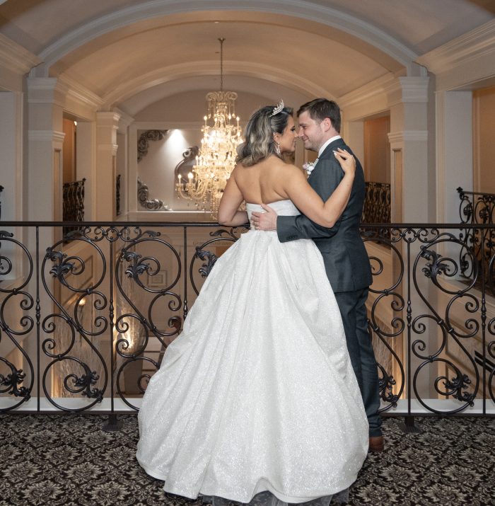 Wedding photo of a bride wearing Anniston by Maggie Sottero with the ballgown skirt bustled, embracing her groom on a balcony beside an ornate iron railing and chandelier.