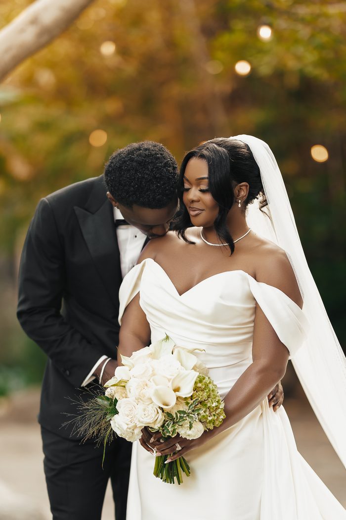 Giselle wedding gown by Sottero and Midgley, an off-the-shoulder sheath dress worn by a bride embraced by her groom in a black tuxedo, holding a white and green bouquet outdoors at golden hour.