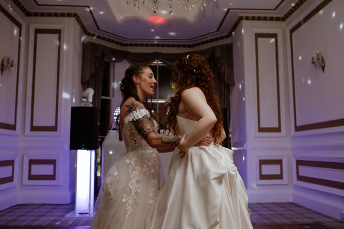 Wedding photo of two brides sharing a first dance in an elegant ballroom, both wearing Maggie Sottero wedding dresses — one a lace ballgown with off-the-shoulder sleeves, the other a strapless satin ballgown.