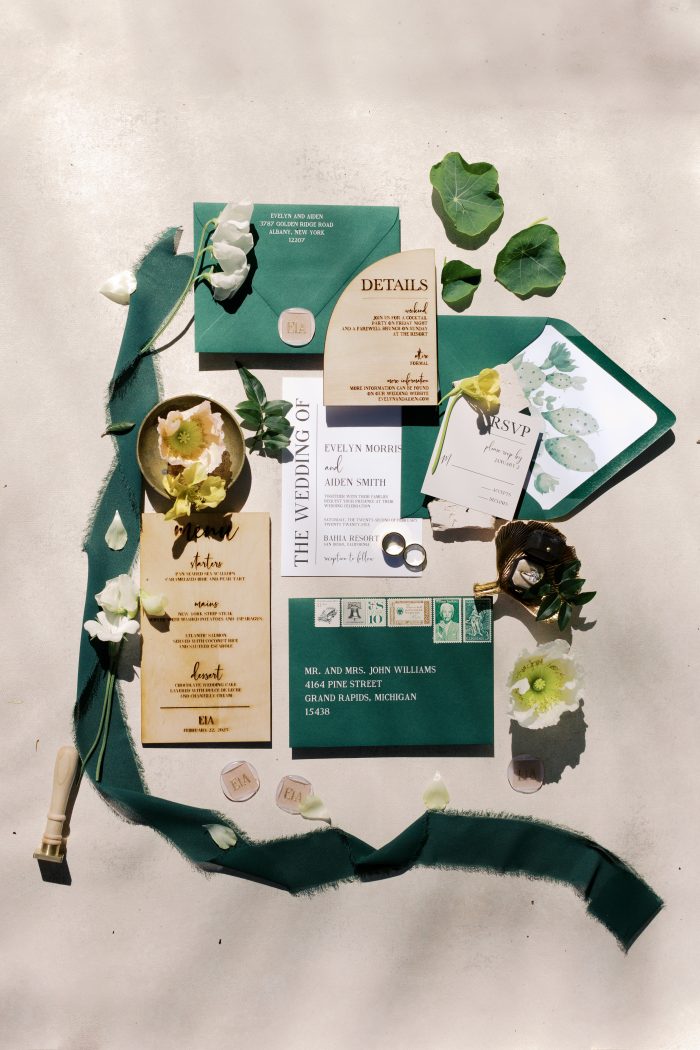 Wedding invitation suite flat lay in a forest green and ivory color palette, with wooden detail cards, envelopes, wax seals, rings, and greenery, photographed by Kellie Hetler.