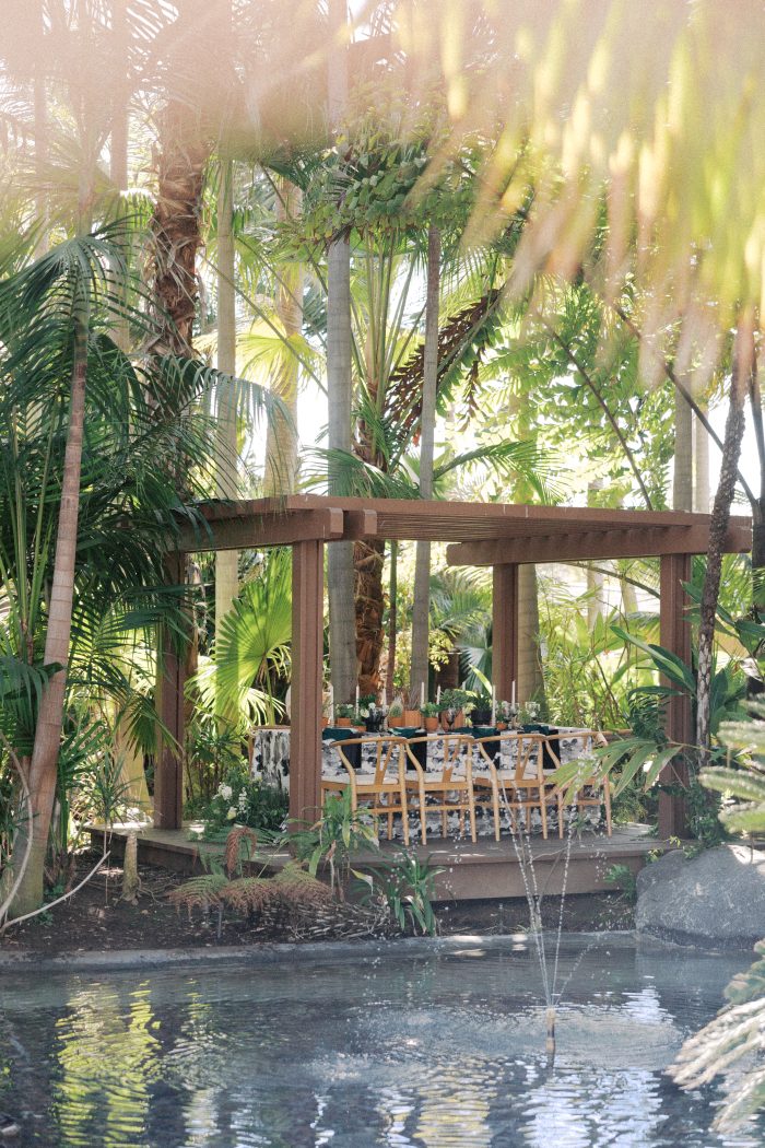 Reception table with wishbone chairs and a black-and-white printed table runner set beneath a wooden pergola surrounded by palm trees and a reflecting pool, photographed by Kellie Hetler.
