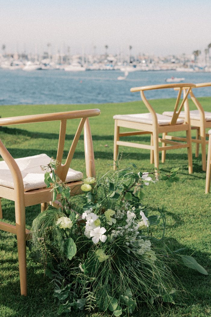 Wishbone ceremony chairs lined with wild greenery and white floral arrangements overlooking a marina, photographed by Kellie Hetler.