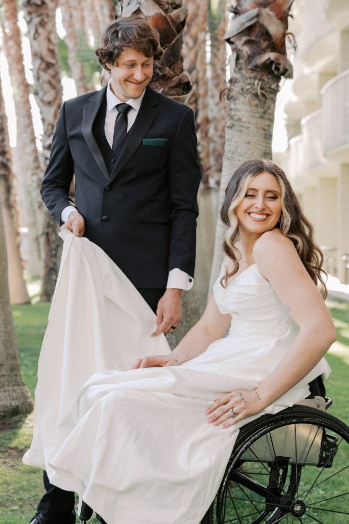 Groom in a black tuxedo helping arrange his bride's Rebecca Ingram wedding gown as she smiles from her wheelchair beneath palm trees, photographed by Kellie Hetler.