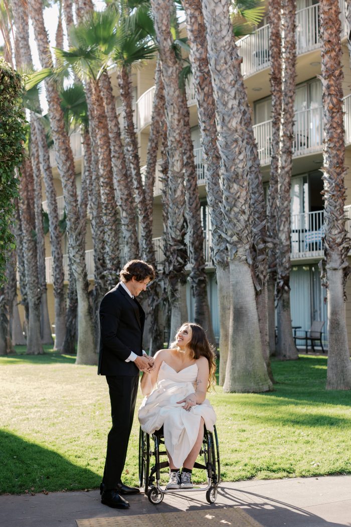 Groom in a black suit holding hands with his bride in her Rebecca Ingram wedding gown along a palm tree-lined path, photographed by Kellie Hetler.