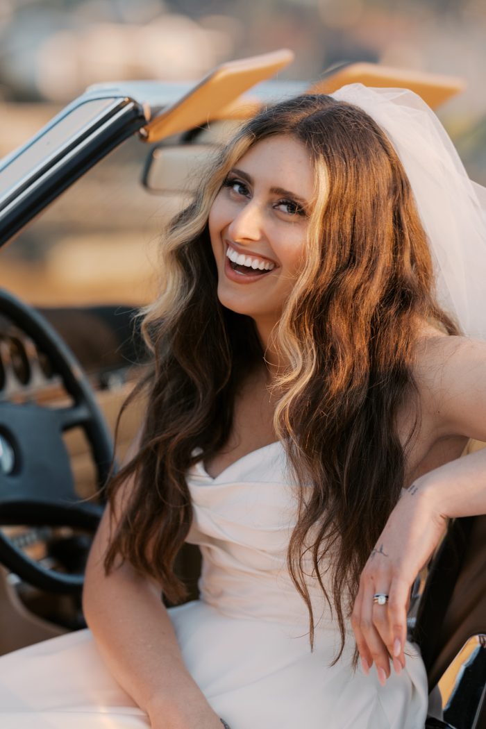 Laughing bride wearing her Rebecca Ingram wedding gown and veil in the back of a vintage convertible, golden hour light on her hair, photographed by Kellie Hetler.
