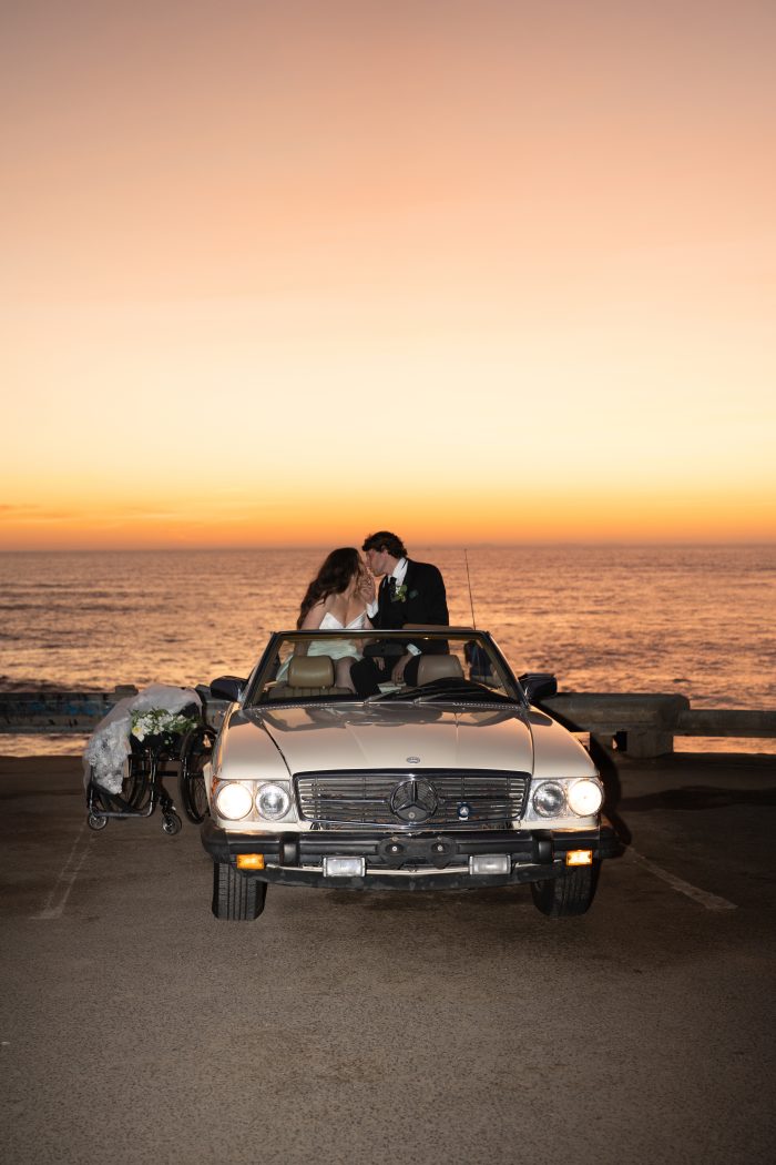 Bride and groom in a vintage convertible sharing a kiss at sunset by the ocean, with the bride's wheelchair parked beside the car, photographed by Kellie Hetler; gown by Rebecca Ingram.