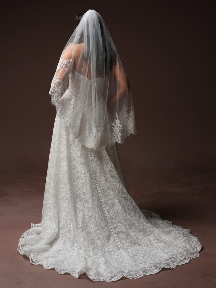 Back view of a curve model wearing the Amante Essentials Edit fingertip veil by Maggie Sottero in ivory with a scalloped lace border, paired with a lace wedding dress featuring a row of covered buttons down the back and a flowing lace train.