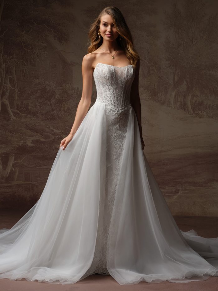 Bride wearing a strapless lace sheath wedding dress with a detachable tulle overskirt train, holding the tulle to reveal the fitted lace gown underneath.