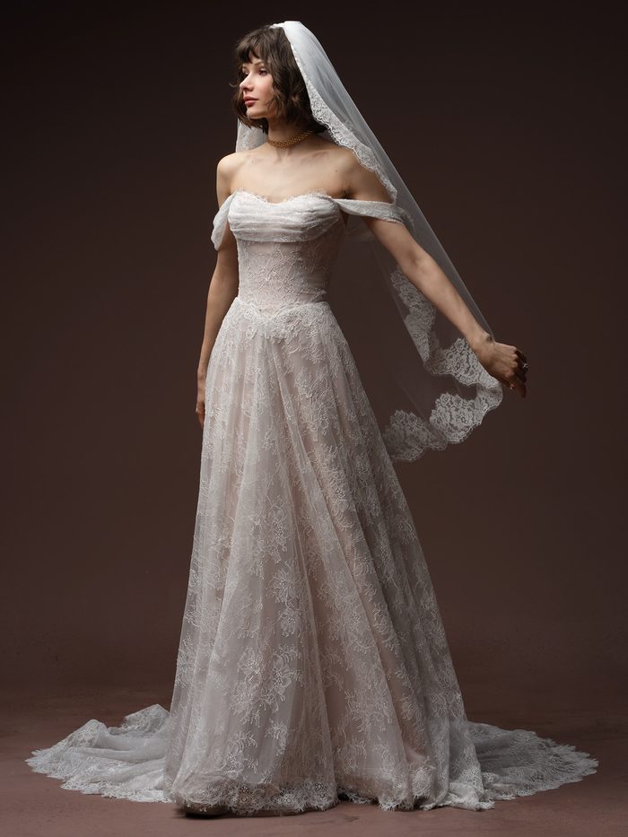 Side back view of a model wearing the Minuet veil from the Essentials Edit by Maggie Sottero, a long lace-edged cathedral veil, paired with a strapless lace wedding dress featuring a scalloped bodice with a row of covered buttons down the back.
