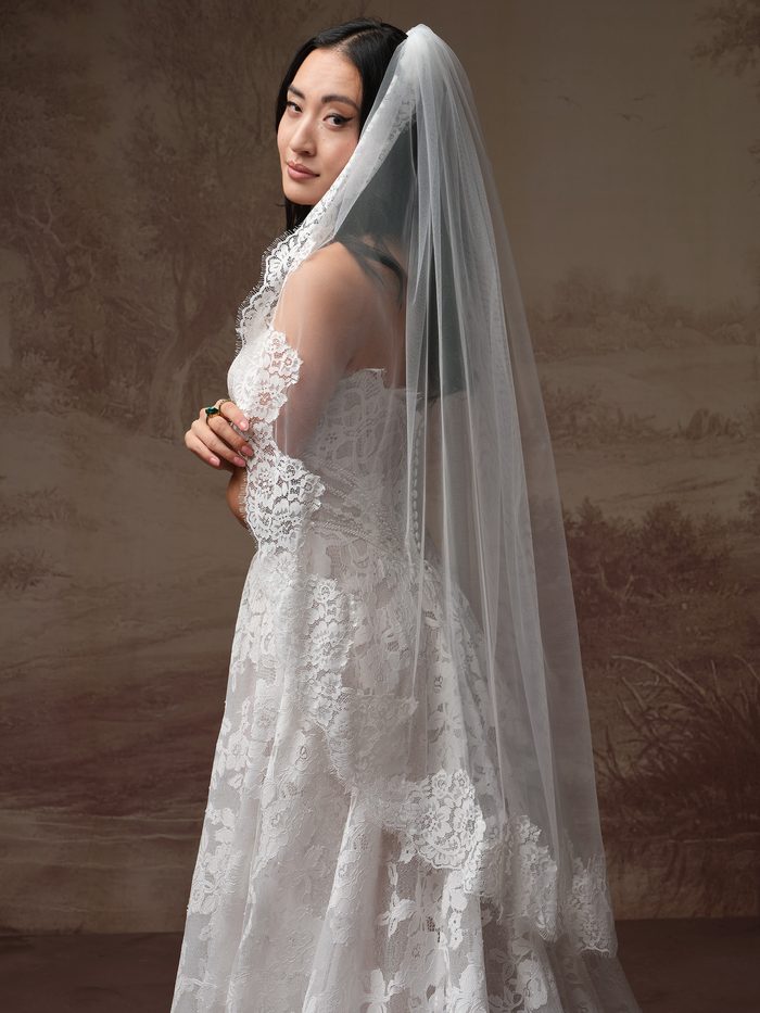 Front view of a model wearing the Minuet veil from the Essentials Edit by Maggie Sottero, a lace-edged veil, paired with an off-the-shoulder lace A-line wedding dress featuring a folded sweetheart neckline and a flowing lace skirt with train.