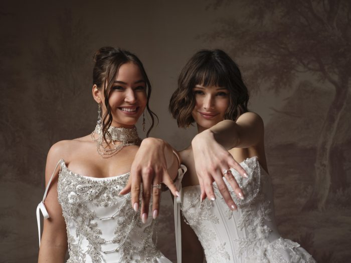 Two smiling brides side by side, both wearing sparkling beaded gowns by Sottero and Midgley — Aelarion and Petra — showing off their engagement rings with hands extended toward the camera.