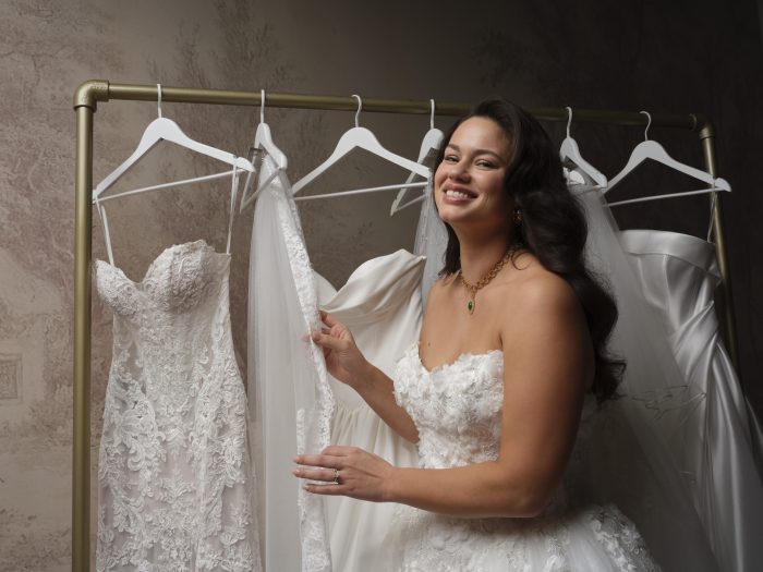Smiling bride wearing the strapless floral 3D lace Benton gown by Maggie Sottero, standing beside a rack of additional wedding dresses with a green pendant necklace and diamond ring.