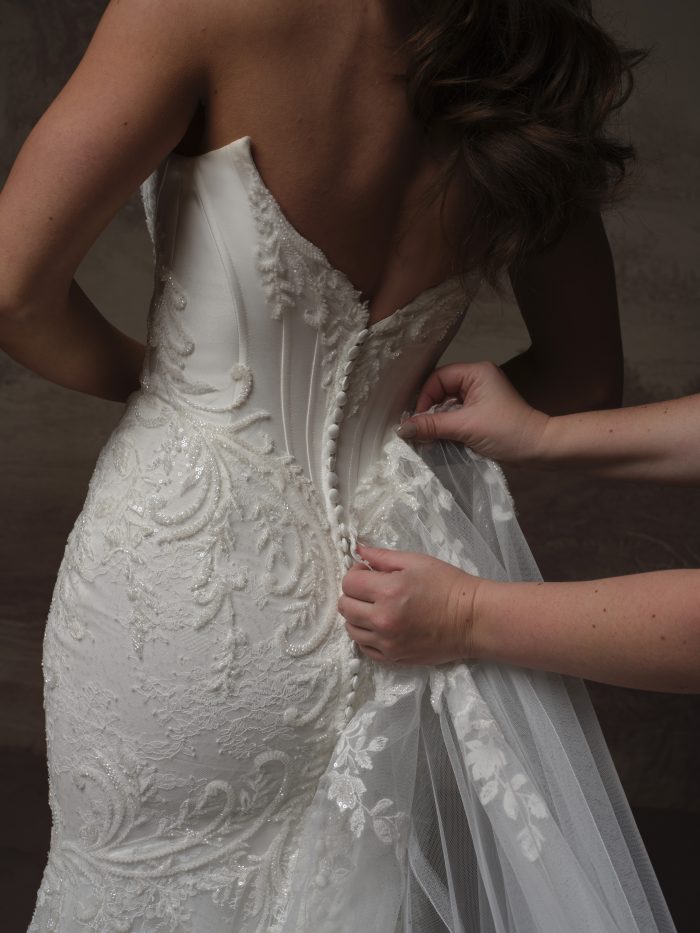 Close-up of the Eirawen gown by Sottero and Midgley from the back, showing the beaded lace-covered button closure and sheer illusion detailing along the spine, with a helper fastening the buttons.