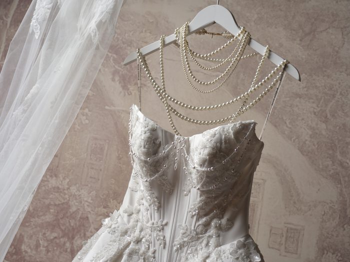 Petra wedding gown by Sottero and Midgley, a strapless lace bodice hanging on a Maggie Sottero branded hanger draped with layered pearl necklaces, with a beaded lace veil beside it.
