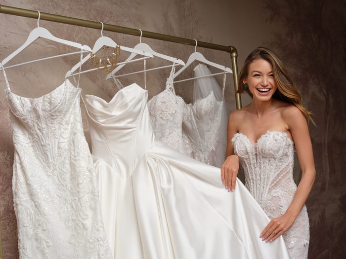 Bride laughing while wearing a beaded strapless Stasia wedding gown by Maggie Sottero, standing beside a rack of additional gowns including beaded lace, satin, and floral lace styles.